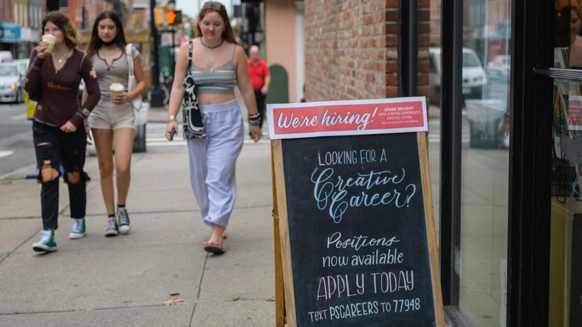 A group of young people walk past a sign that says We’re hiring!