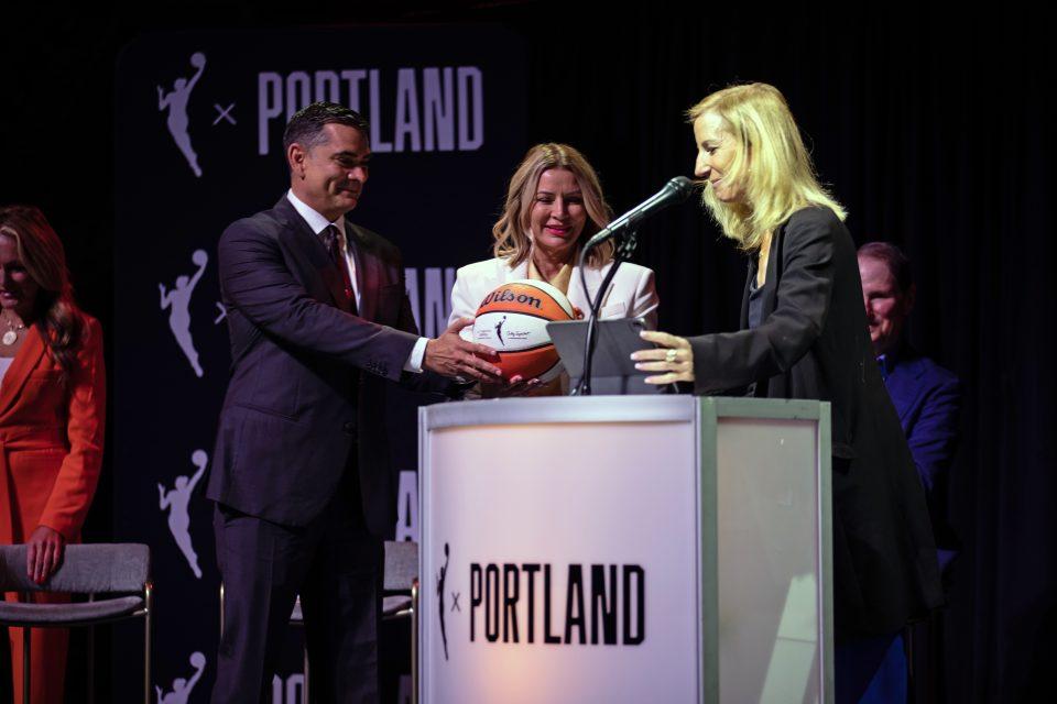 gettyimages-2172257091-e1788559039387 Alex Bhathal and Lisa Bhathal Merage stand next to a podium and accept a basketball from Cathy Engelbert.